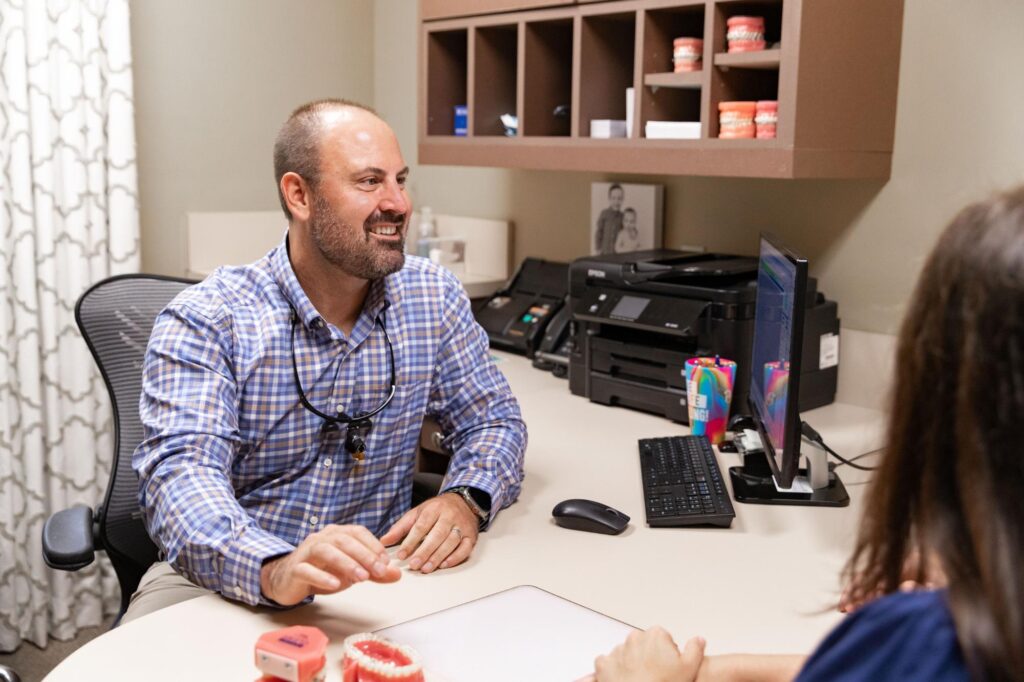 Dr. Ryan Cox in a blue plaid shirt, sitting at a workstation in the clinic and smiling during a patient appointment at Ryan Cox Orthodontics in Rome, GA